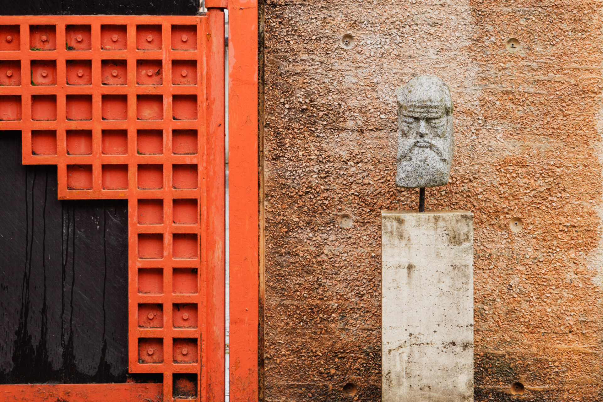 Scultura "Stone Head" - Una scultura in pietra con una testa su un piedistallo contro una parete marrone testurizzata è accanto a una grande porta a traliccio rossa e nera. La scena combina elementi rustici e geometrici, creando un sorprendente contrasto visivo.