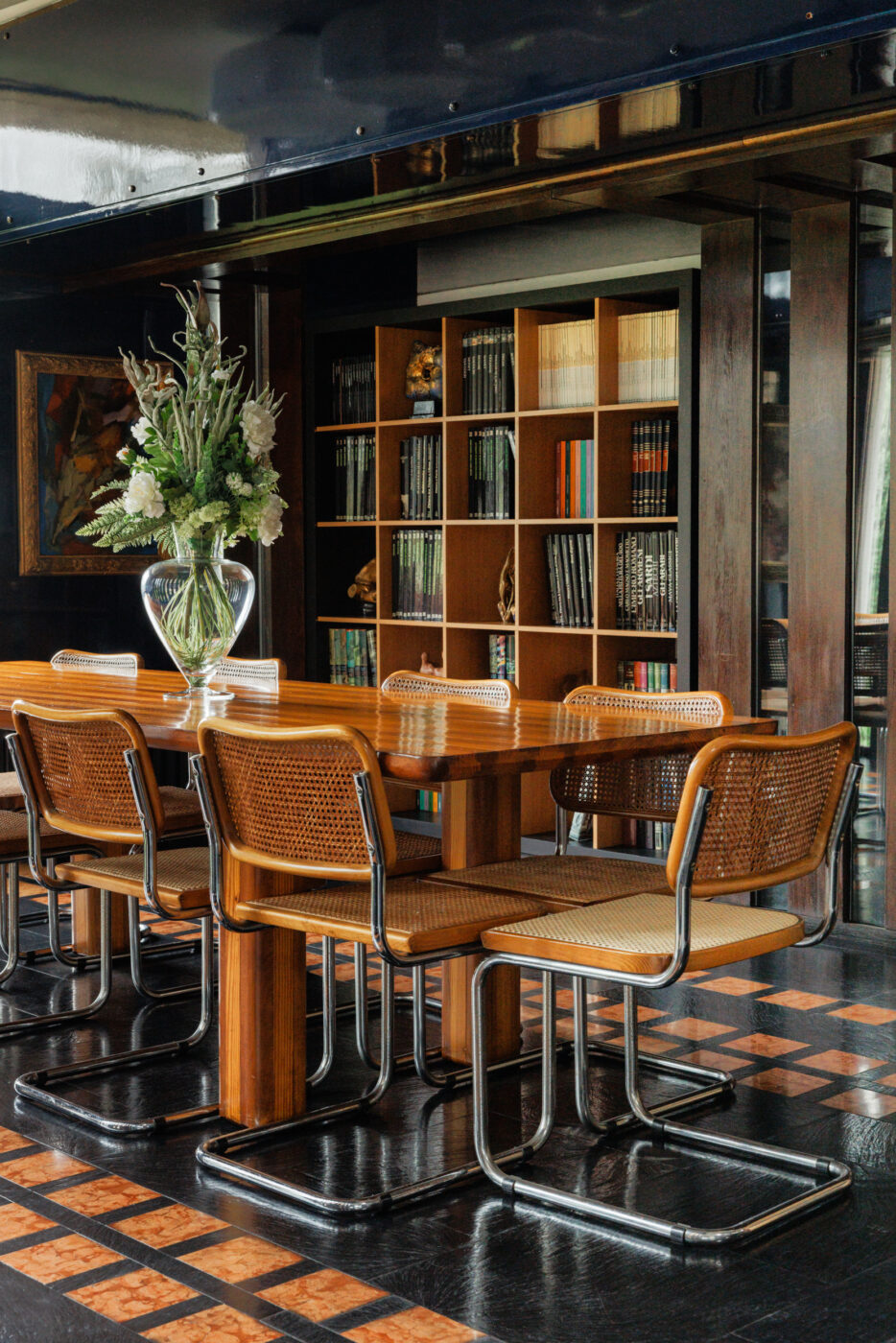 Modern Dining Room" - Una moderna sala da pranzo con un tavolo di legno, sei sedie di vimini e una grande composizione floreale in un vaso di vetro. Librerie piene di libri e decorazioni fiancheggiano la parete posteriore. La stanza è caratterizzata da un pavimento scuro con accenti a motivo intrecciato.
