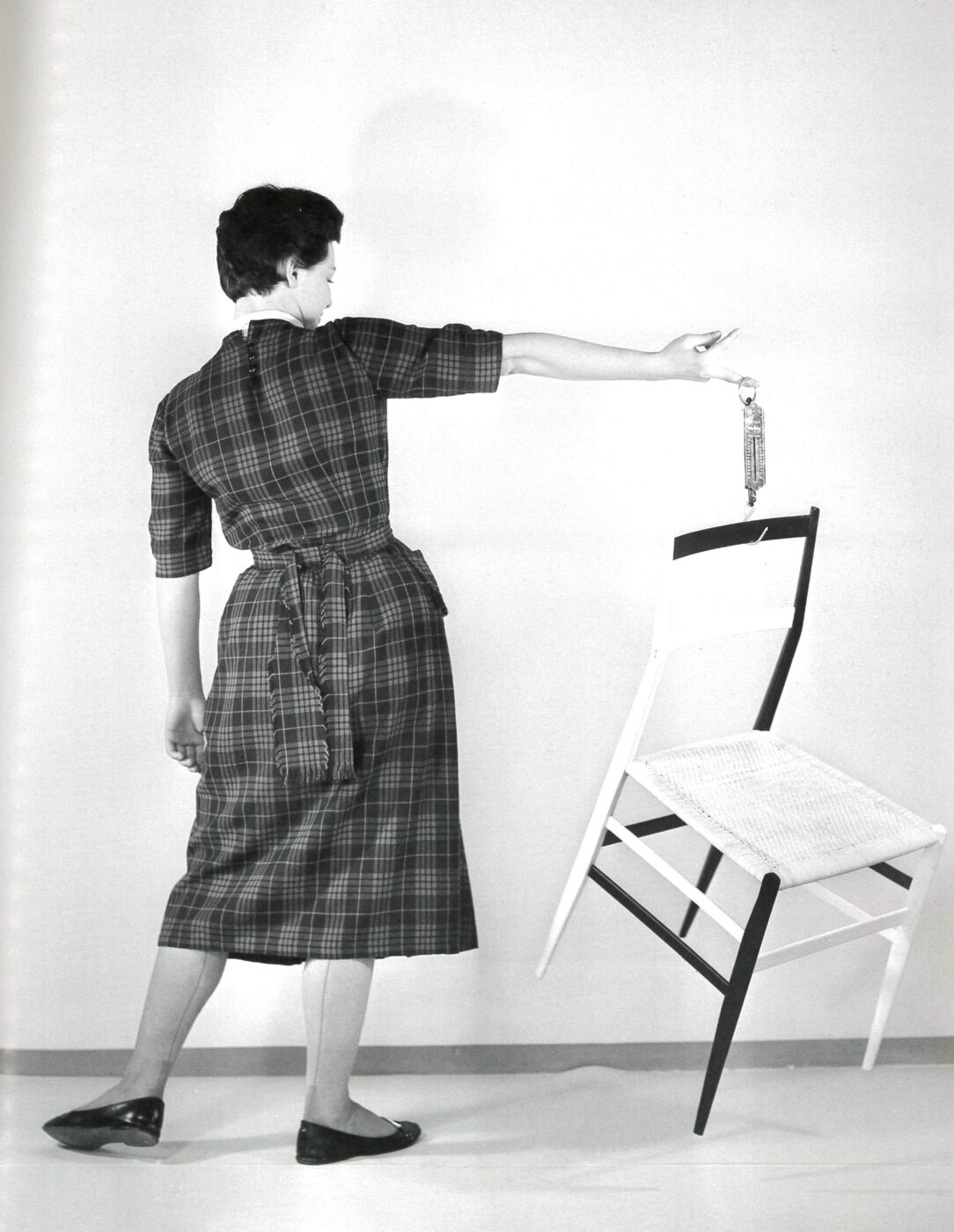 Gio Ponti's Superleggera chair - A person in a checkered dress stands on one foot, balancing on the edge of a white chair with one foot and holding a small bottle in one hand. The background is plain, creating a minimalist and artistic scene.