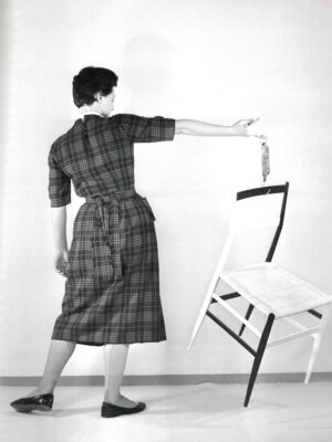 Gio Ponti's Superleggera chair - A person in a checkered dress stands on one foot, balancing on the edge of a white chair with one foot and holding a small bottle in one hand. The background is plain, creating a minimalist and artistic scene.