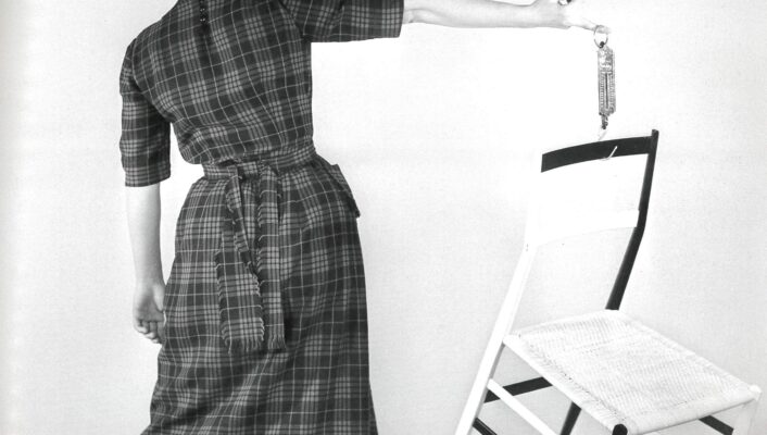 Gio Ponti's Superleggera chair - A person in a checkered dress stands on one foot, balancing on the edge of a white chair with one foot and holding a small bottle in one hand. The background is plain, creating a minimalist and artistic scene.