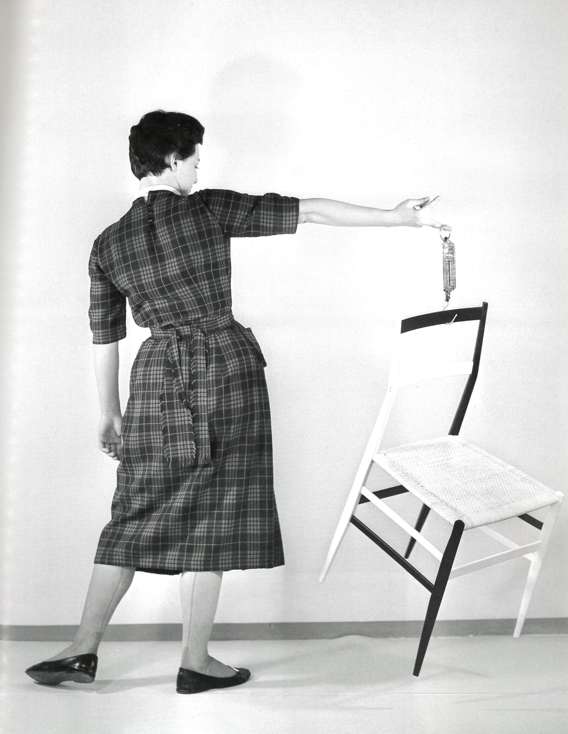Gio Ponti's Superleggera chair - A person in a checkered dress stands on one foot, balancing on the edge of a white chair with one foot and holding a small bottle in one hand. The background is plain, creating a minimalist and artistic scene.