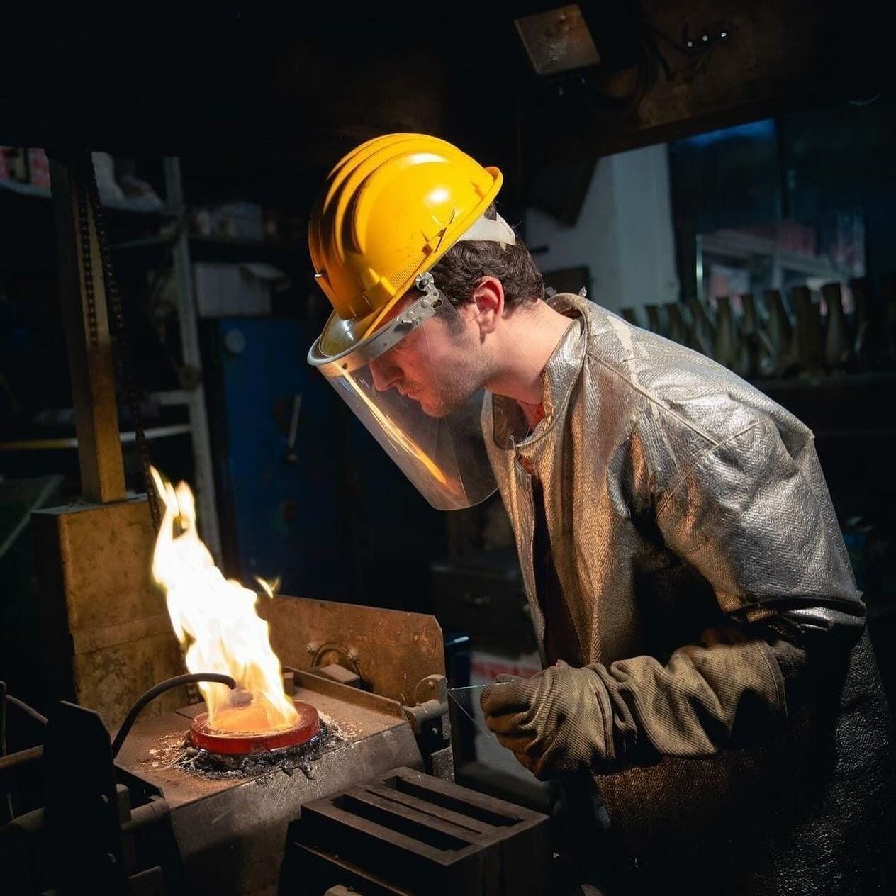 Pampaloni - A person wearing a yellow hard hat, face shield, and protective gloves is closely observing a fiery metalworking process in an industrial setting. They are dressed in a heat-resistant silver jacket, standing near equipment amid dim lighting.