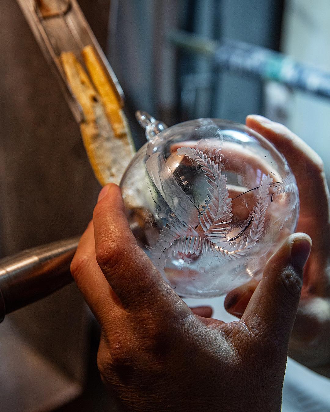Moleria Locchi - Hands hold a clear glass object with intricate floral engravings. A tool rests on the object, suggesting craftsmanship and precision. The background is blurred, focusing on the detailed etchings.