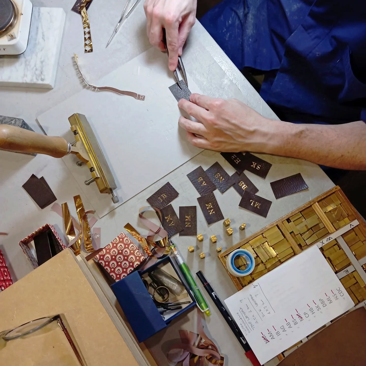 Atelier GK - Person working at a desk with tools and materials for bookbinding. There are pieces of leather, gold letters, glue, a hammer, and a calendar. The person uses tweezers to handle small items.