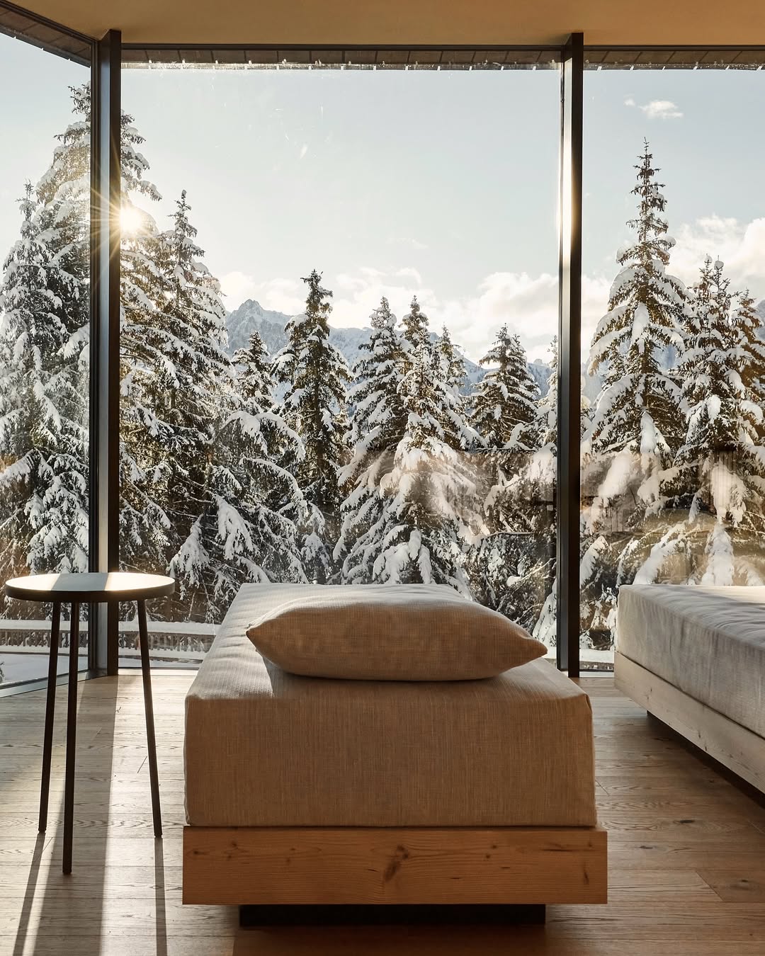 Forestis – A cozy room with floor-to-ceiling windows offers a stunning view of snow-covered pine trees and mountains. The room features a simple wooden bed, a gray cushion, and a small round table, with sunlight streaming through. If theres a specific image title youd like to include at the beginning of this alt text, please provide it!.