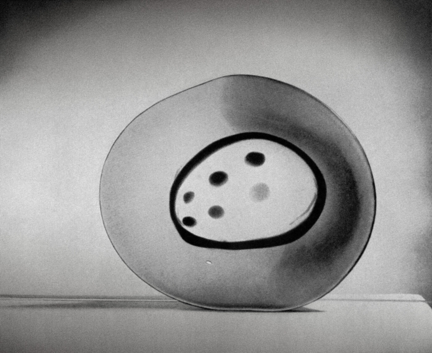 A grayscale abstract sculpture featuring a circular, plate-like form with an oval center and dotted patterns. The sculpture sits on a flat surface with soft lighting and a plain background.