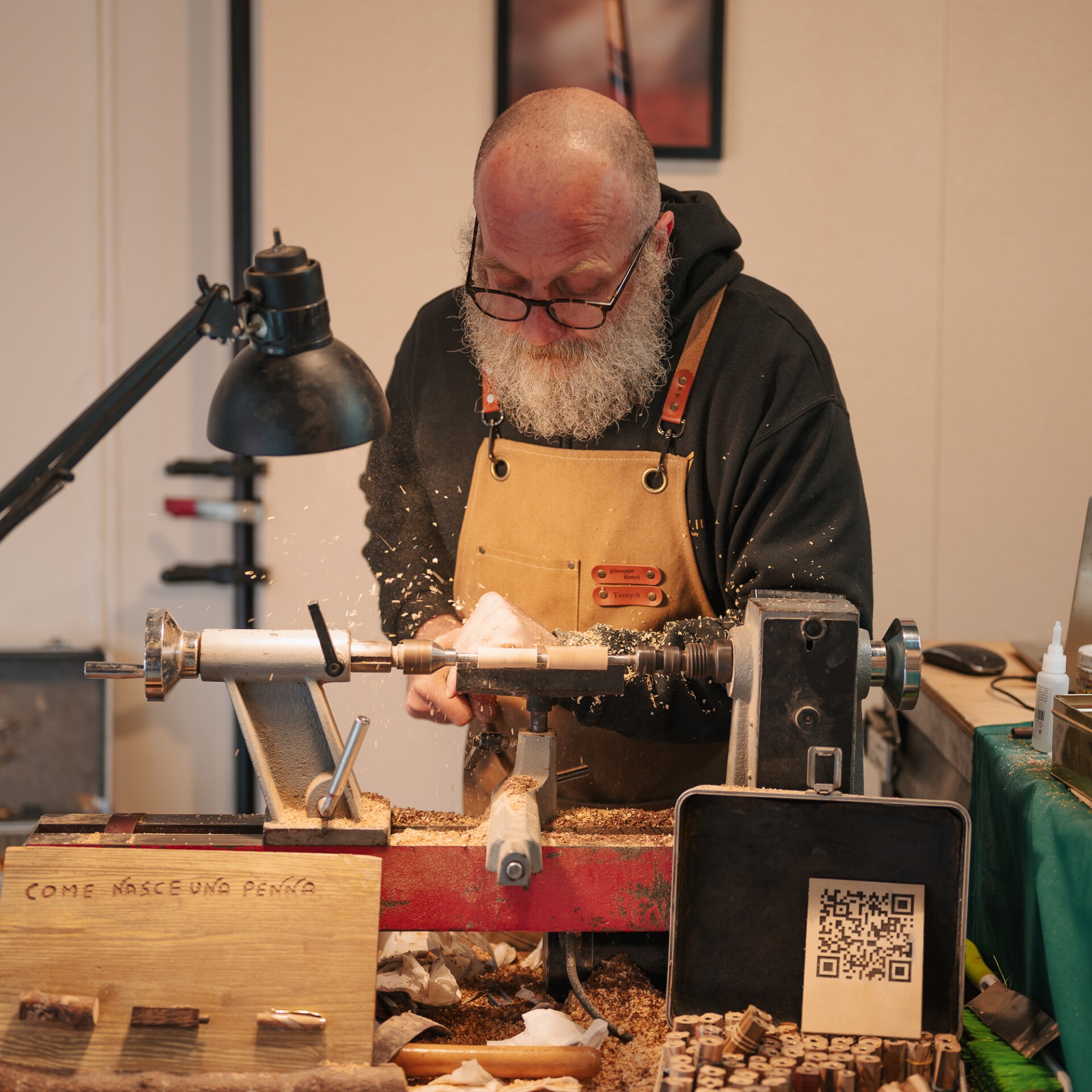Woodworking at Night - Alt Text: A bearded man wearing glasses and an apron is using a lathe to shape wood, surrounded by wood shavings. Theres a lamp above him, a sign with Italian text, and a QR code on a tablet nearby.