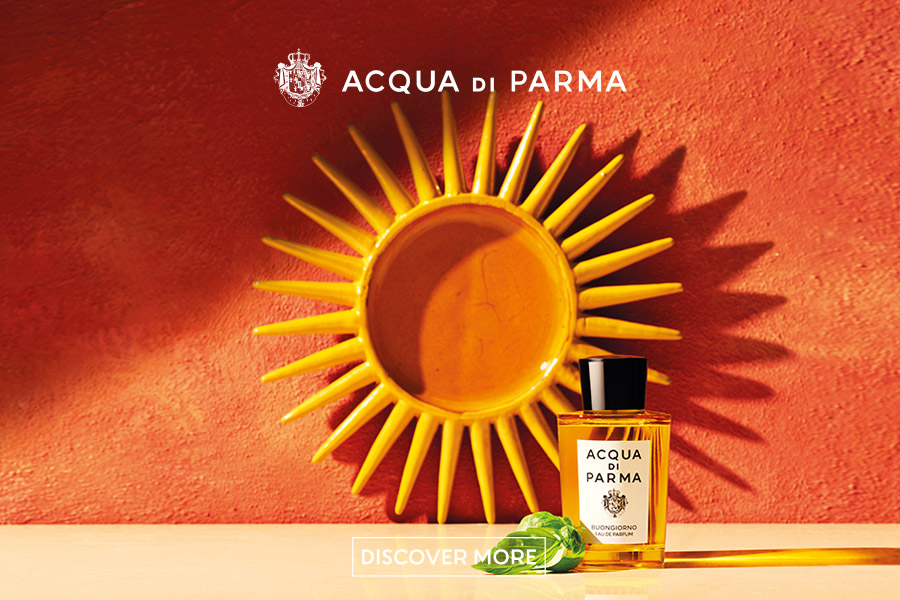 Acqua di Parma Perfume" - Una bottiglia di profumo Acqua di Parma è posizionata davanti a una decorazione murale a forma di sole su uno sfondo rosso strutturato. Il testo "ACQUA DI PARMA" e "DISCOVER MORE" appare sopra e sotto la bottiglia.