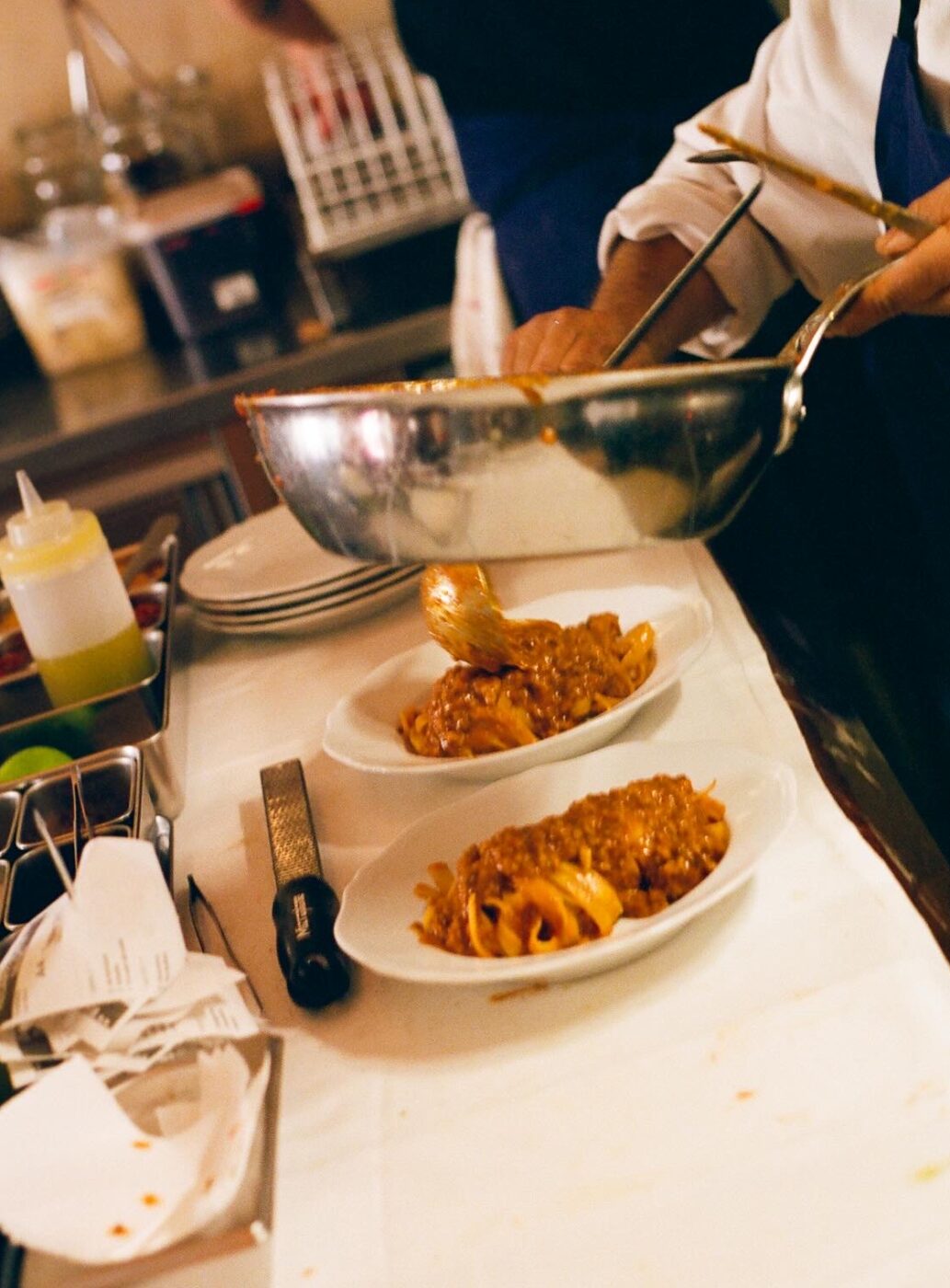 Localino, Paris– A chef is serving pasta onto plates in a kitchen. The pasta is coated in a red sauce and is placed on a counter with various kitchen utensils and ingredients nearby.