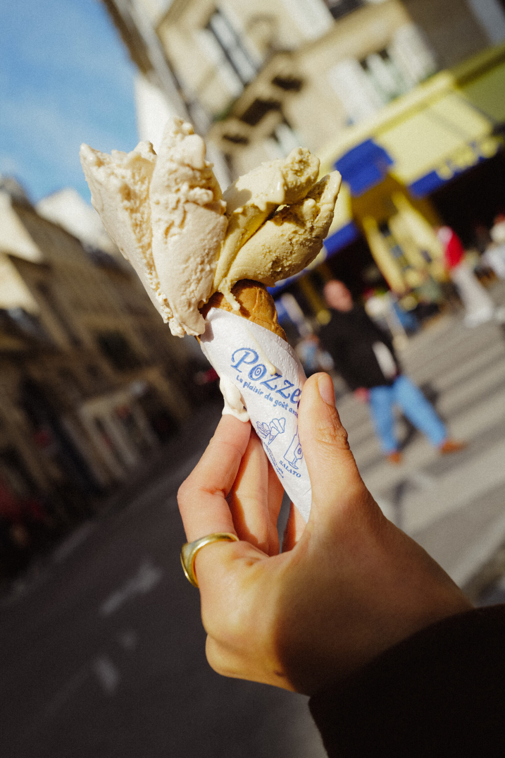 Street Gelato Delight" - Mano che tiene un gelato cremoso in un cono avvolto in carta con il marchio, con una scena di strada sullo sfondo con bar e pedoni, sotto un limpido cielo azzurro.