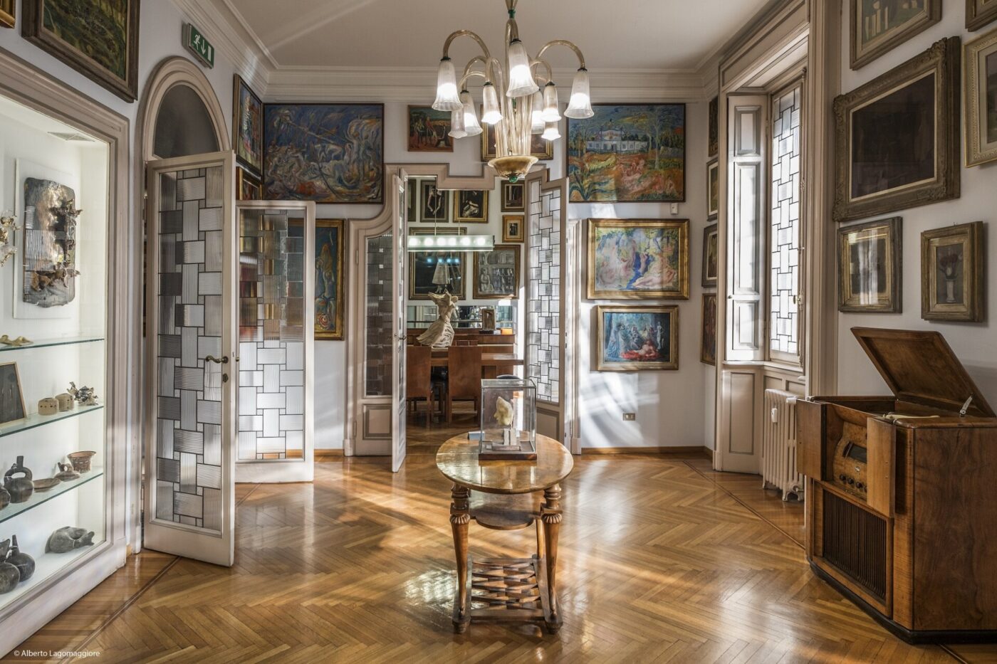 Elegant Room - Elegante camera con pavimento in parquet, decorata con opere d'arte e sculture. Un tavolo rotondo è sistemato al centro sotto un lampadario classico, circondato da ampie finestre e porte aperte con pannelli in vetro. Un lettore musicale d'epoca è visibile sulla destra.