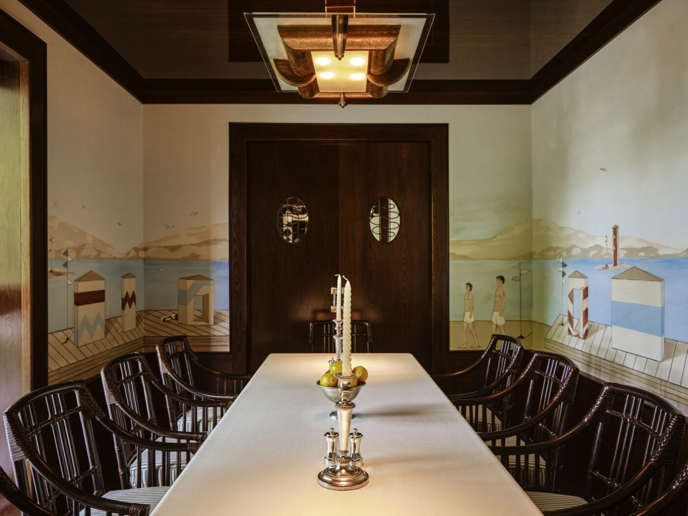 Elegante sala da pranzo" - Elegante sala da pranzo con un lungo tavolo coperto da una tovaglia bianca, un candelabro centrale e circondato da sedie in legno scuro. Le pareti sono decorate con murales di scene di mare e una moderna lampada a sospensione è appesa sopra.