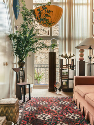 Navarin, Pari - A stylish living room featuring a peach sofa and a patterned red rug in front of a marble fireplace. The space is illuminated by large windows, while leafy plants and eclectic artwork bring added charm. A large lamp is suspended from the ceiling, and a glass door opens to reveal a small balcony.