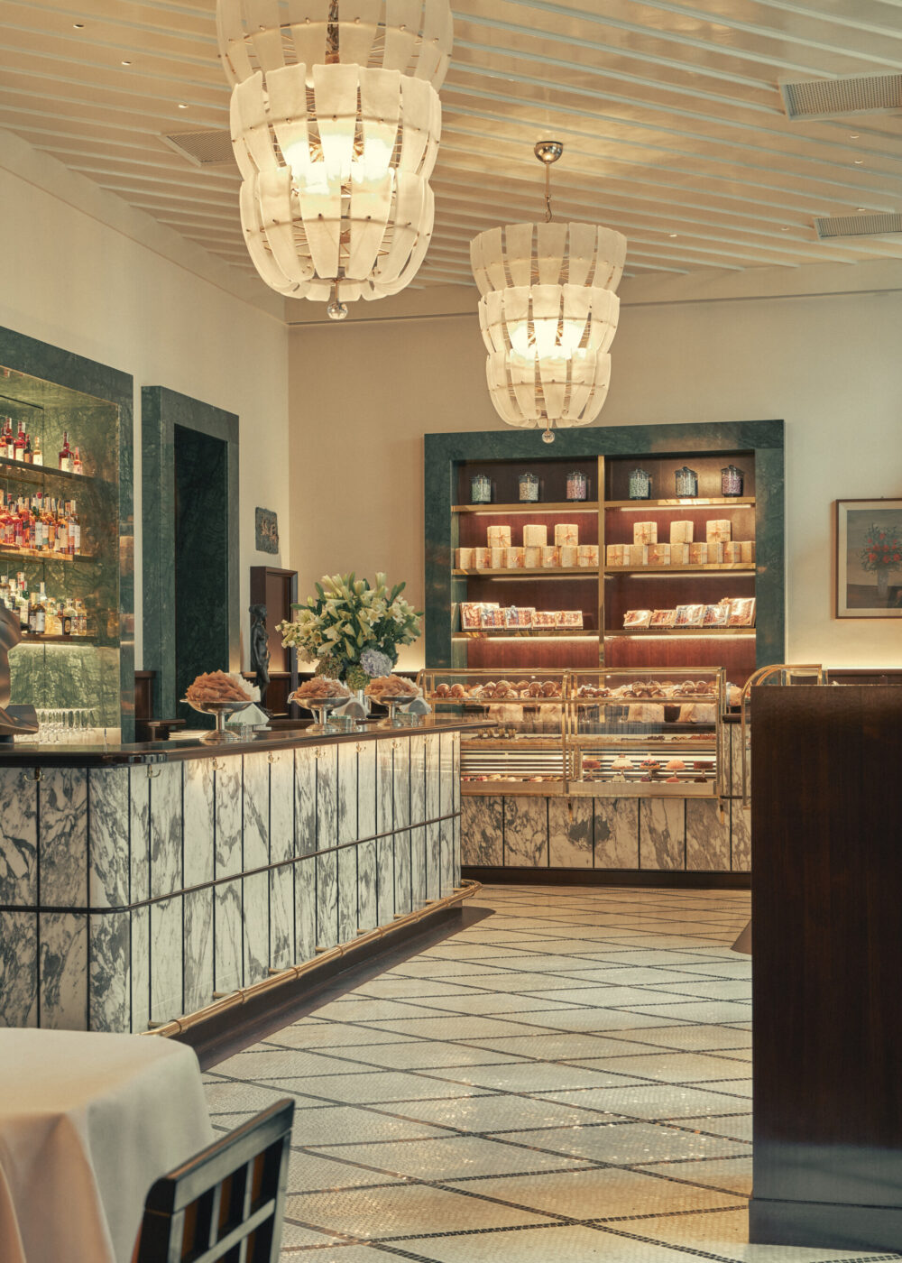 Elegante interno di una pasticceria - Elegante interno di una pasticceria con banconi in marmo, scaffali espositivi pieni e grandi lampadari. Gli scaffali espongono una varietà di prodotti confezionati, mentre fiori freschi adornano il bancone. Il pavimento presenta un motivo geometrico di piastrelle.
