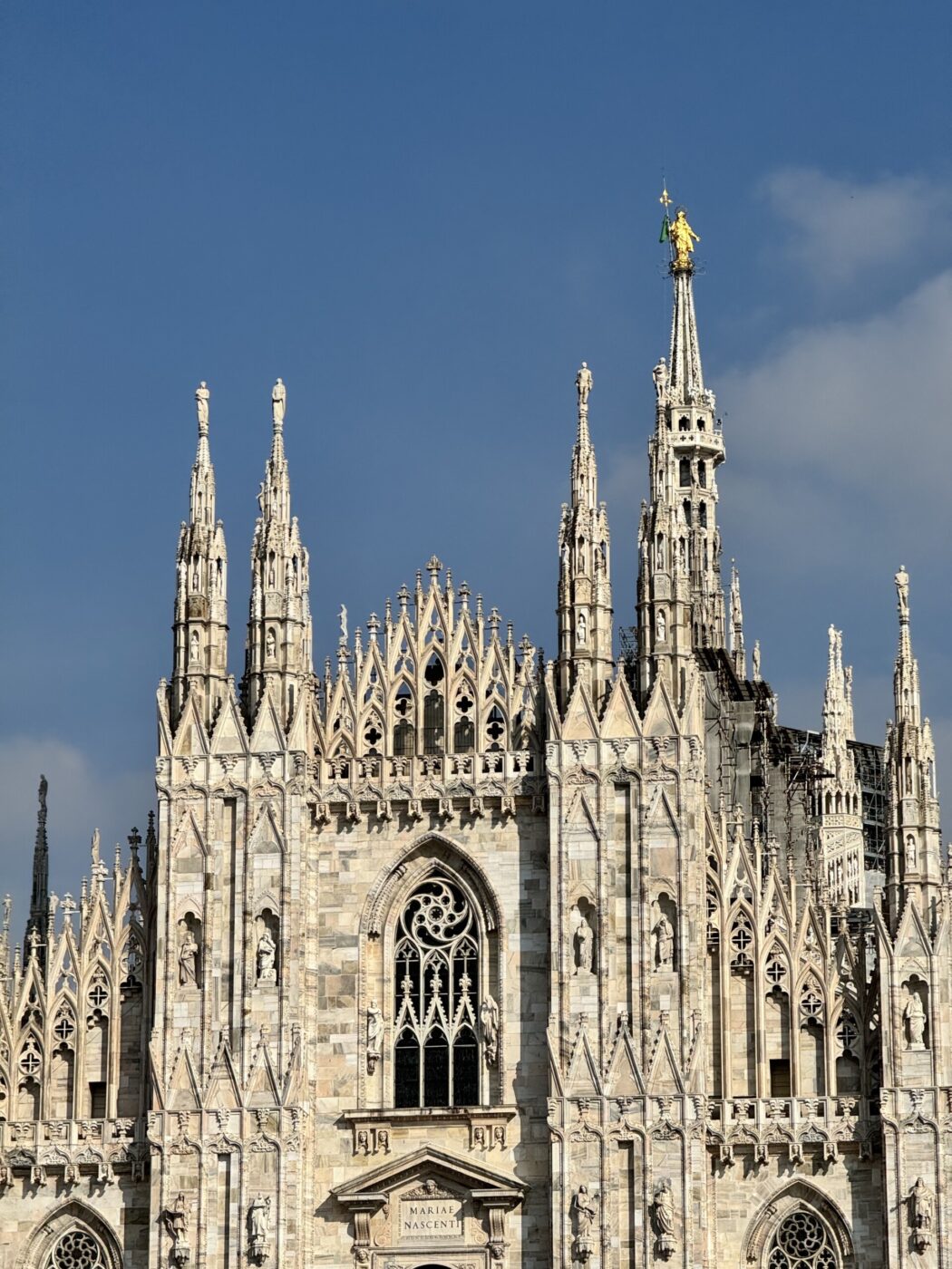 Facciata del Duomo di Milano" - L'immagine mostra la facciata ornata del Duomo di Milano sotto un cielo azzurro e limpido. L'intricata architettura gotica presenta guglie appuntite, sculture decorative e una grande finestra centrale. Una statua dorata incorona la guglia più alta.