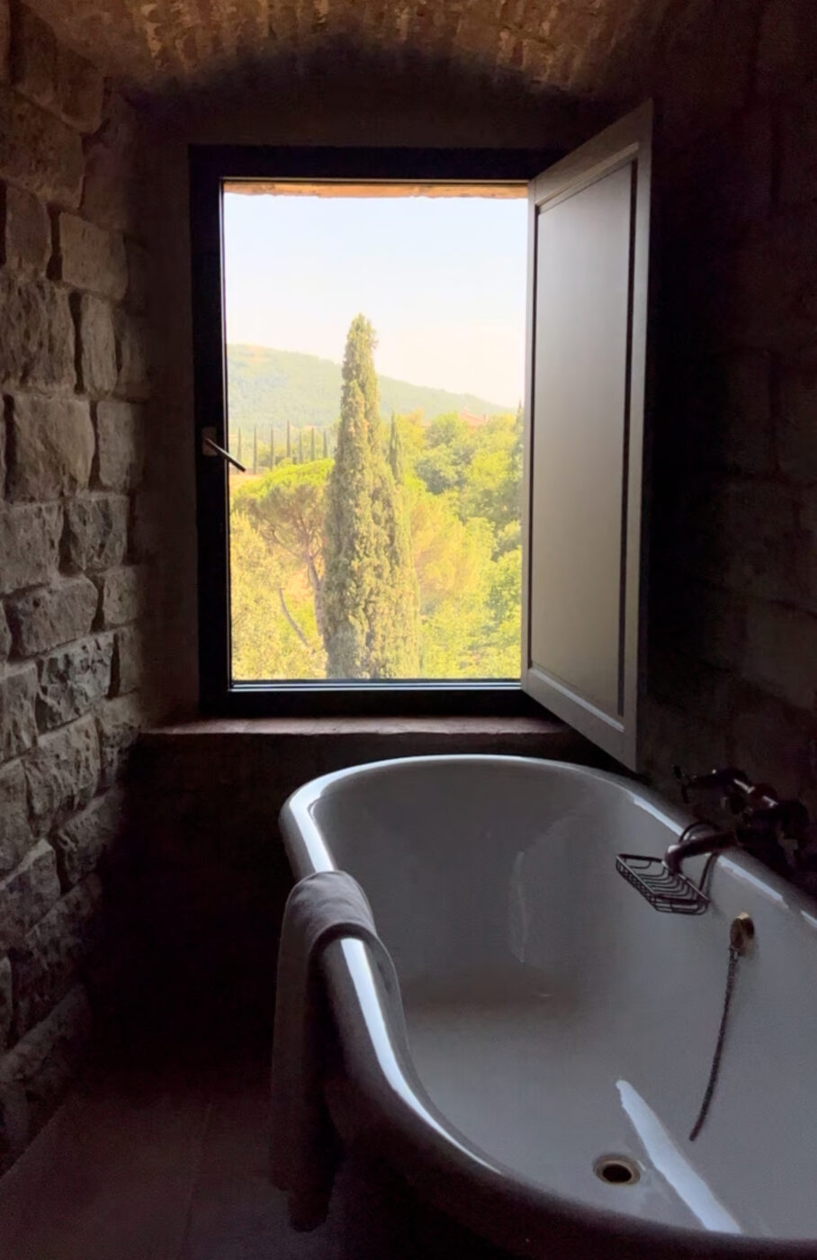 Rustic Bathroom with Scenic View - Alt Text: A rustic stone bathroom with a white clawfoot bathtub is shown. A towel hangs over the tubs edge. An open window reveals a scenic view of lush green hills and trees under a clear sky, casting soft light into the room.