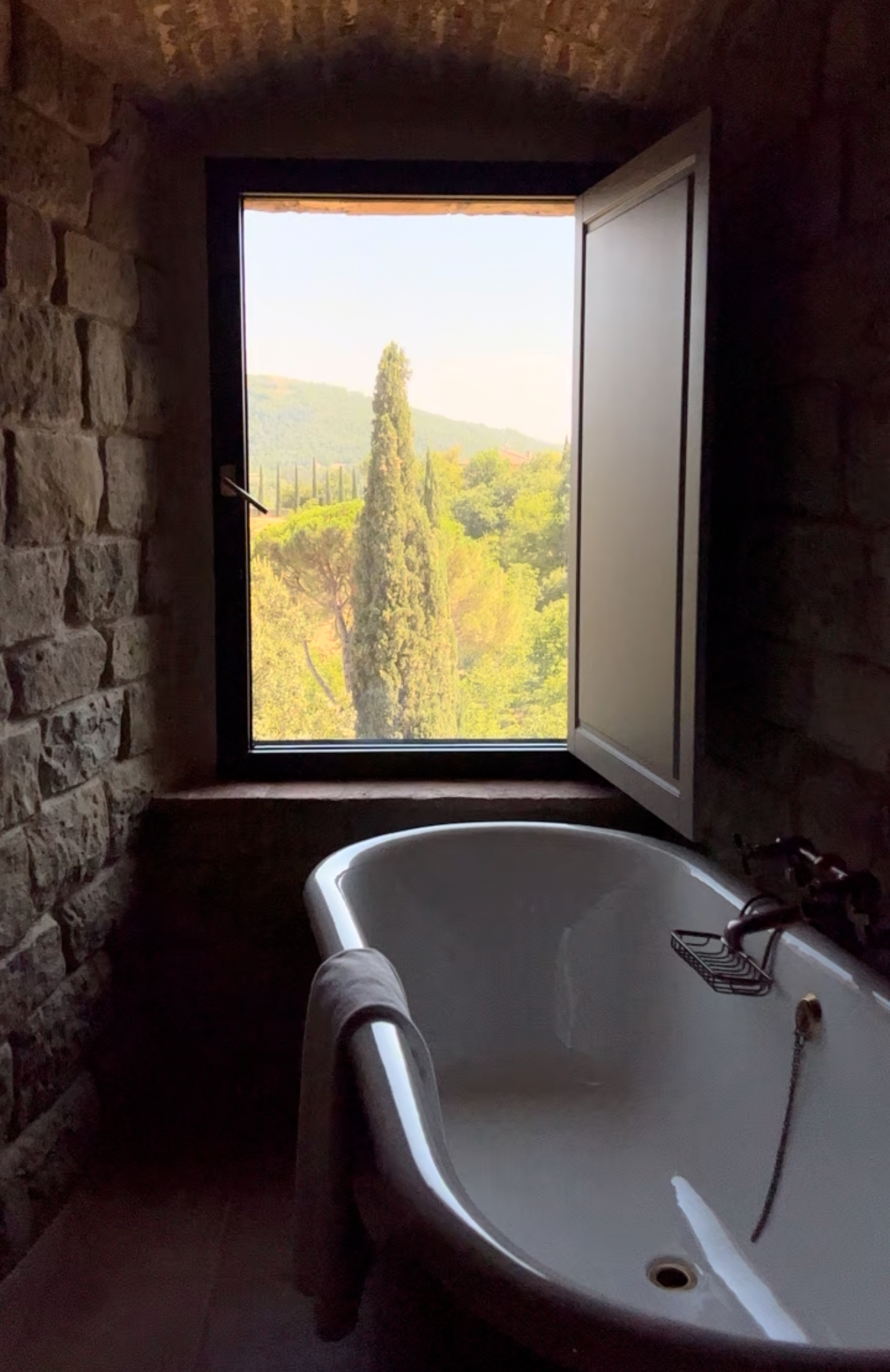 Rustic Bathroom with Scenic View - Alt Text: A rustic stone bathroom with a white clawfoot bathtub is shown. A towel hangs over the tubs edge. An open window reveals a scenic view of lush green hills and trees under a clear sky, casting soft light into the room.
