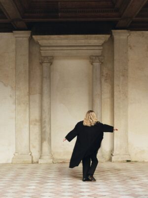 It seems you are asking for the alt text with the image title included at the beginning. However, there is no image title provided in your prompt. If you have an image title, simply insert it before A person in your description like this: [Image Title] - A person with long blonde hair, wearing a long black coat, stands with arms slightly outstretched in front of a worn architectural backdrop with columns. The floor is checkered, and the atmosphere is serene and contemplative. If you provide an actual image title, I can help further customize it for you!.
