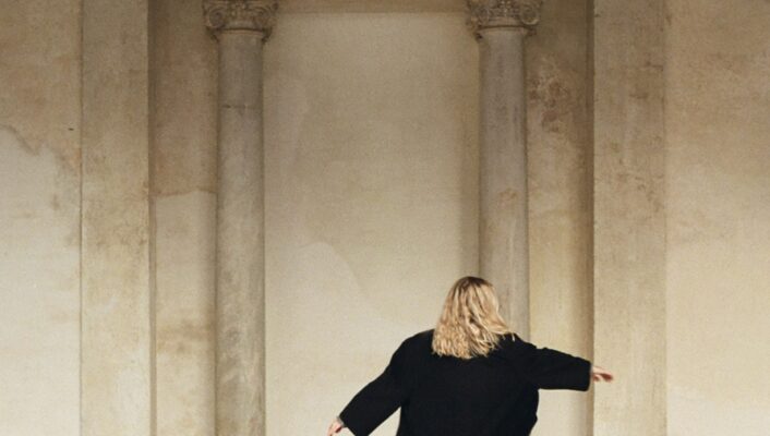 It seems you are asking for the alt text with the image title included at the beginning. However, there is no image title provided in your prompt. If you have an image title, simply insert it before A person in your description like this: [Image Title] - A person with long blonde hair, wearing a long black coat, stands with arms slightly outstretched in front of a worn architectural backdrop with columns. The floor is checkered, and the atmosphere is serene and contemplative. If you provide an actual image title, I can help further customize it for you!.