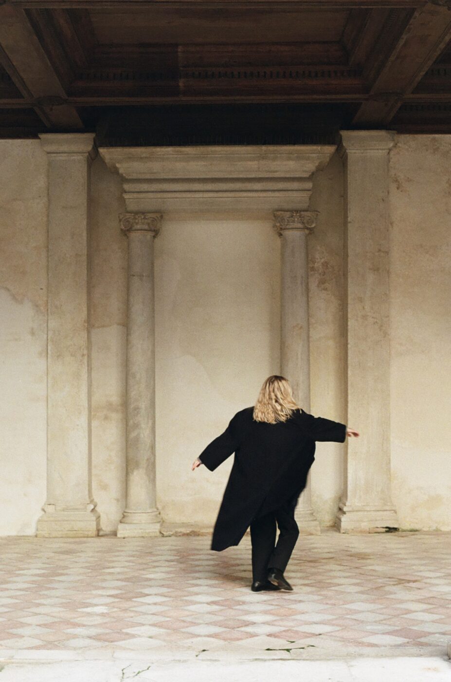 Sembra che non ci sia un titolo dell'immagine fornito nella tua descrizione. Pertanto, il testo alternativo sarebbe: "Persona con lunghi capelli biondi in un cappotto nero in piedi in una spaziosa stanza con architettura d'epoca, tra cui alte colonne e un pavimento a scacchi. Sono rivolti altrove, e sembrano danzare o girare leggermente.