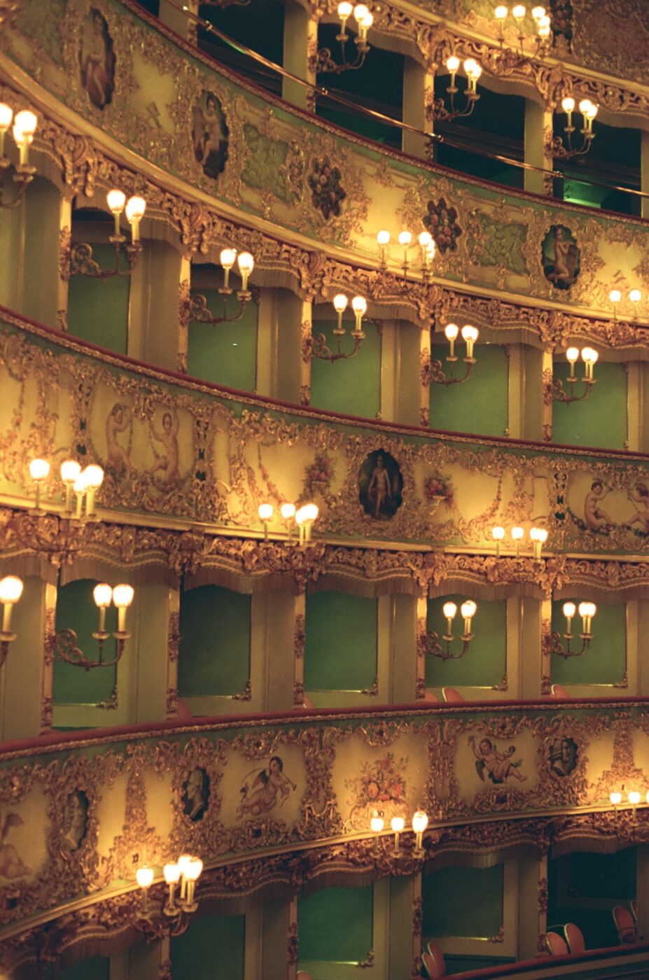 Eleganti interni teatrali" - Alt Text: Eleganti interni teatrali con molteplici balconi ornati, ornati da modanature decorative e luci soffuse e calde provenienti da numerose lampadine luminose. I ricchi dettagli e i sedili lussuosi evocano un senso di grandezza classica.