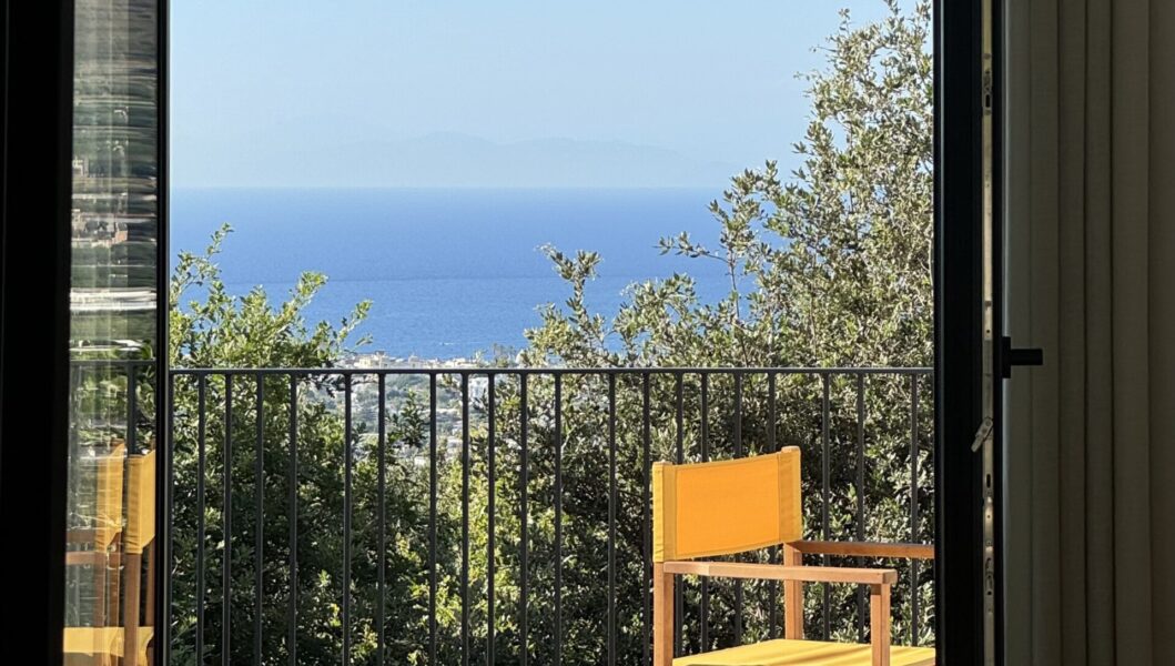 Casa Via Costa - A sunny patio with a simple wooden chair facing a railing. Beyond the railing, lush greenery overlooks a vast expanse of blue ocean under a clear sky. A door frame partially encloses the view.