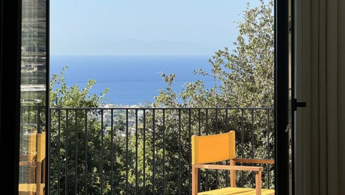 Casa Via Costa - A sunny patio with a simple wooden chair facing a railing. Beyond the railing, lush greenery overlooks a vast expanse of blue ocean under a clear sky. A door frame partially encloses the view.