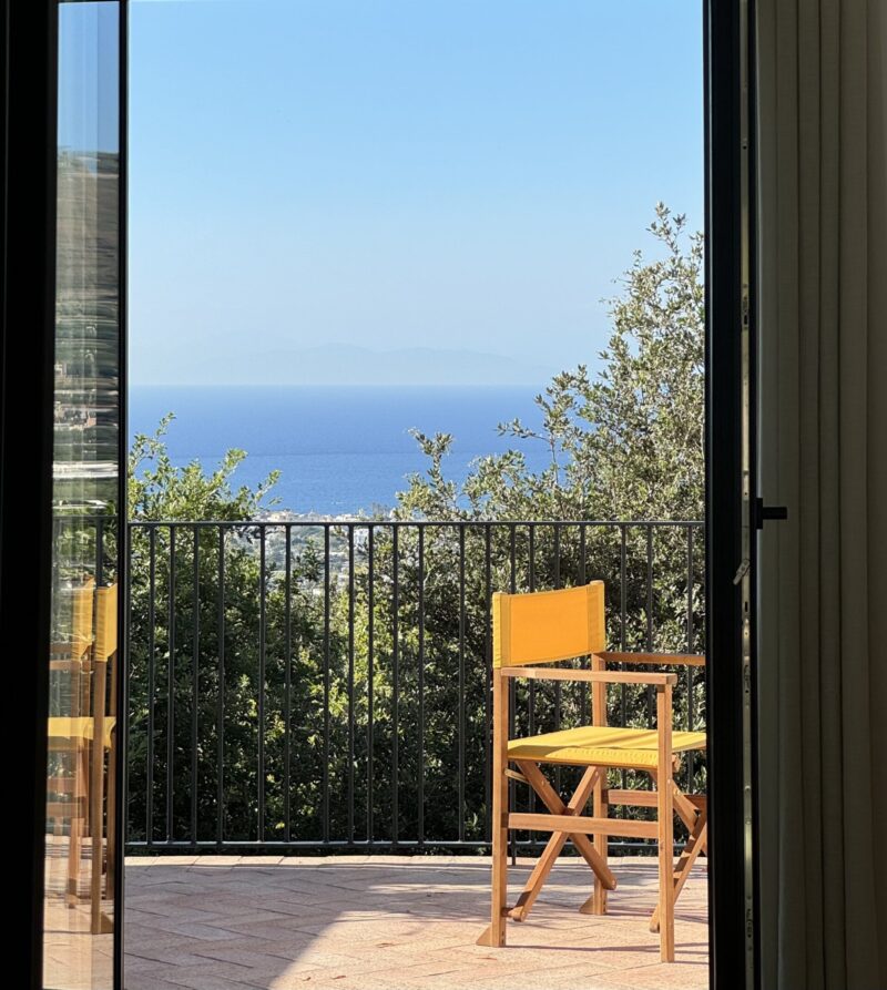 Casa Via Costa - A sunny patio with a simple wooden chair facing a railing. Beyond the railing, lush greenery overlooks a vast expanse of blue ocean under a clear sky. A door frame partially encloses the view.