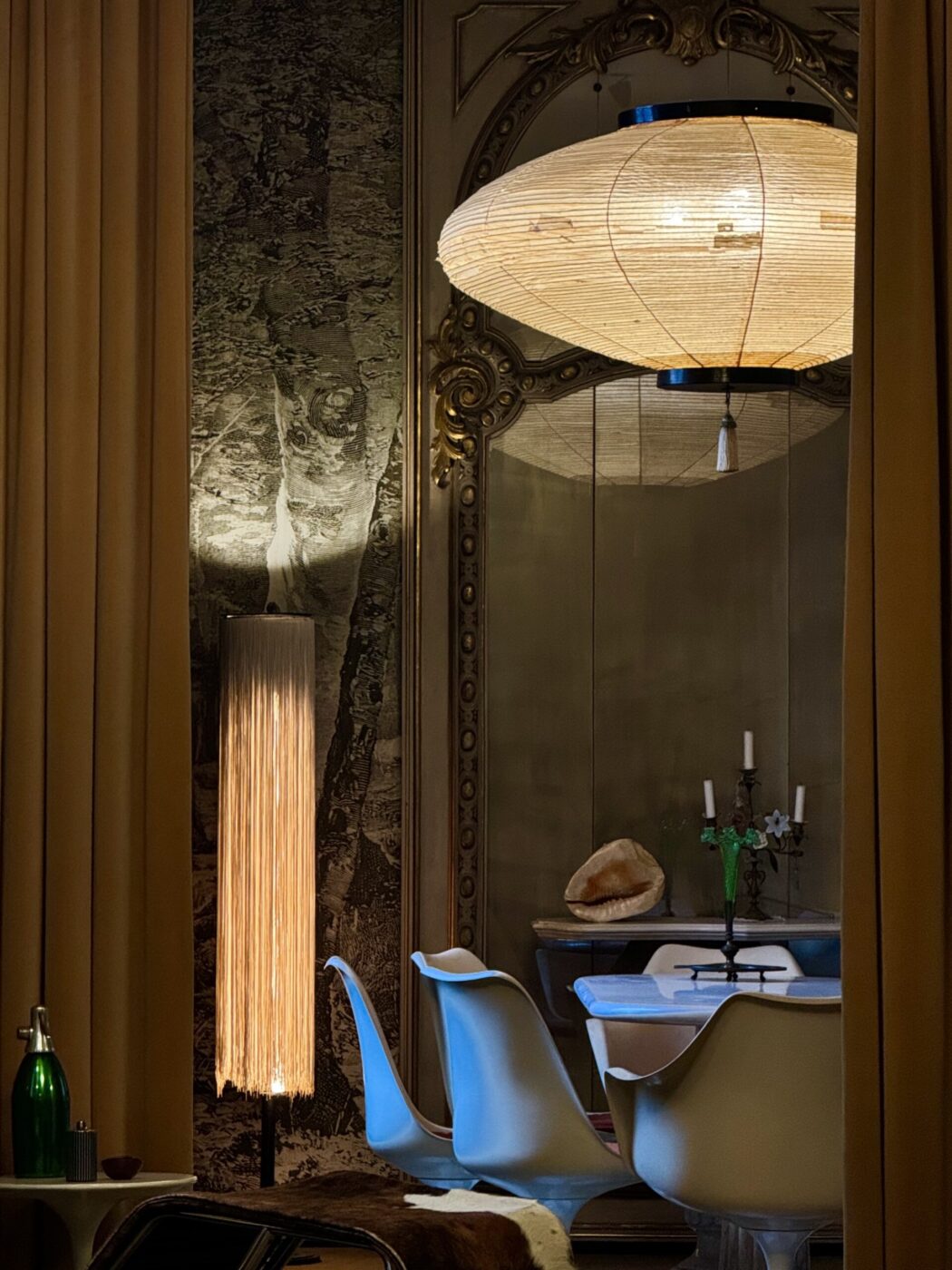 Interni eleganti con una lampada da terra alta, sedie bianche moderne, un tavolo rotondo e una grande lampada sospesa. Tende beige incorniciano la scena e una parete strutturata aggiunge profondità. Una bottiglia verde e oggetti decorativi adornano lo spazio.