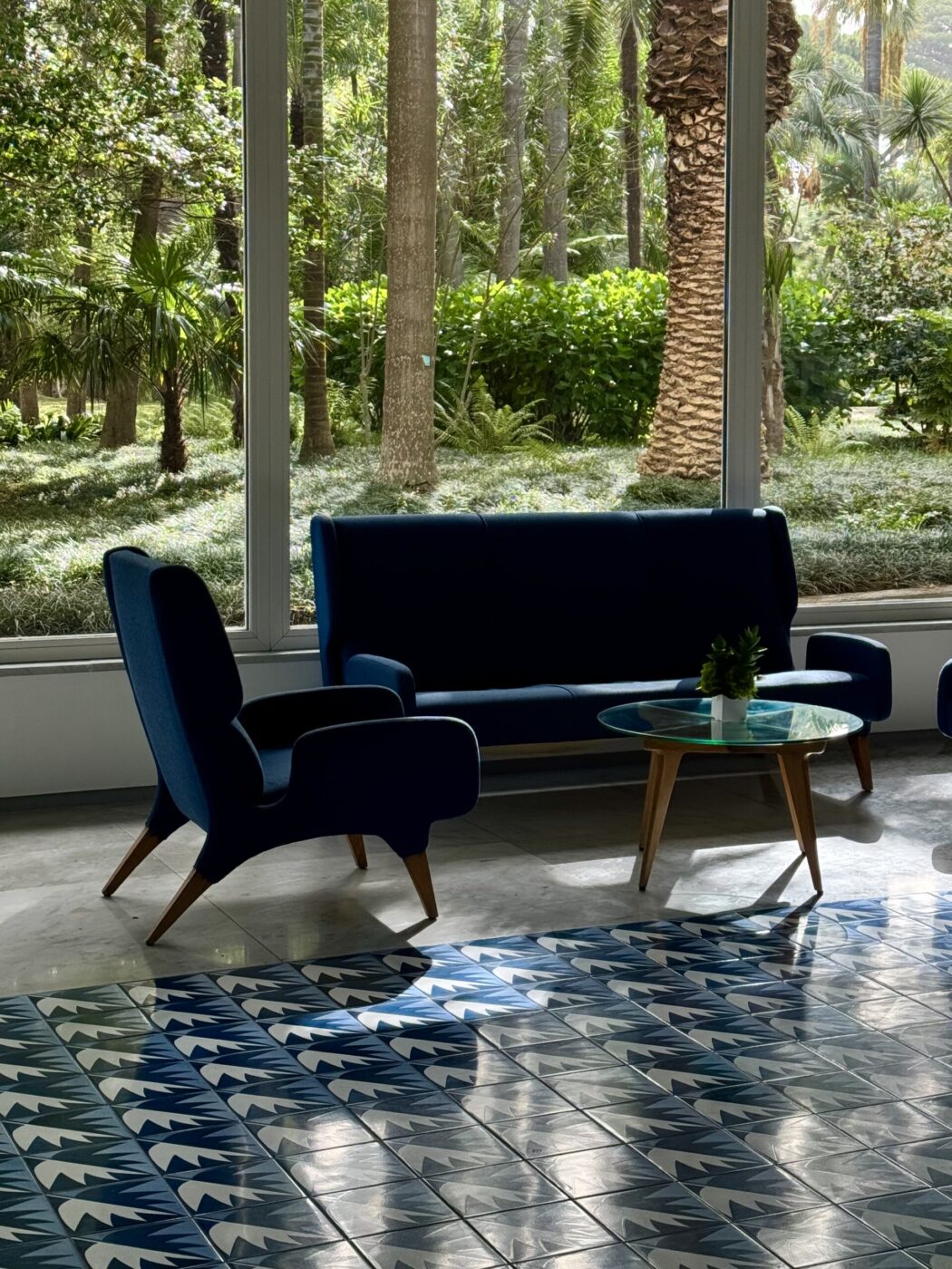 [Titolo dell'immagine] - Una moderna area lounge con un divano blu e due poltrone abbinate attorno a un tavolino da caffè in vetro. La stanza ha grandi finestre che rivelano una vegetazione lussureggiante all'esterno. Piastrelle blu e bianche con motivi geometrici ricoprono il pavimento.