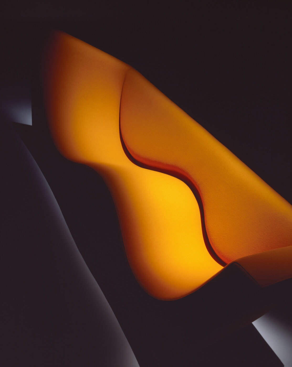 Three Sofa de Luxe, designed by Jasper Morrison for the Cappellini photo by Santi Caleca - Close-up of a modern, sculptural piece of art with flowing curves and smooth, interwoven shapes. The artwork is illuminated with a warm, orange and yellow gradient, creating a glowing effect against a dark background.