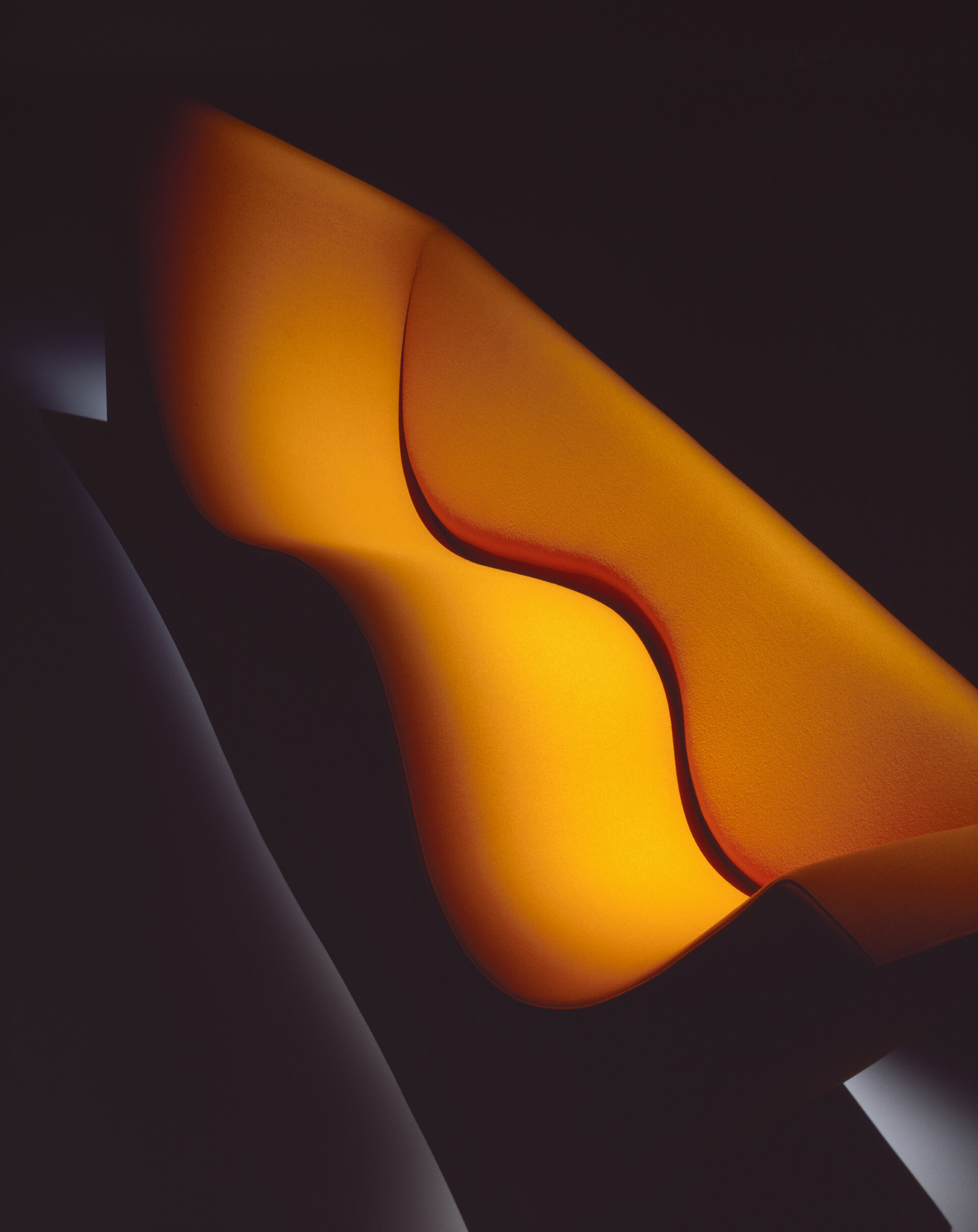 Three Sofa de Luxe, designed by Jasper Morrison for the Cappellini photo by Santi Caleca - Close-up of a modern, sculptural piece of art with flowing curves and smooth, interwoven shapes. The artwork is illuminated with a warm, orange and yellow gradient, creating a glowing effect against a dark background.