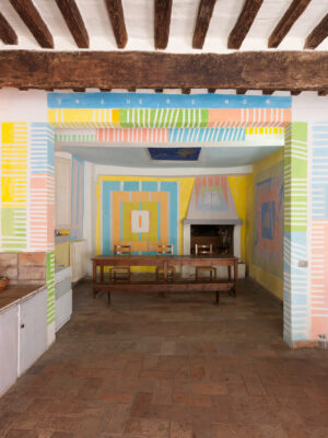 The Kitchen of Casa Dipinta - Colorful room with geometric patterns on walls and beams. Main wall features blue, orange, and pink shapes around a fireplace. Wooden beams contrast with white ceiling. ONE HERE NOW painted in blue. Table and chairs in center, tile floor.