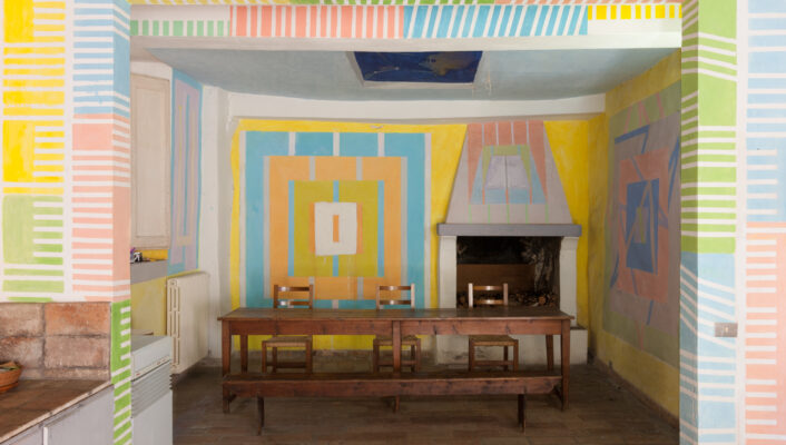 The Kitchen of Casa Dipinta - Colorful room with geometric patterns on walls and beams. Main wall features blue, orange, and pink shapes around a fireplace. Wooden beams contrast with white ceiling. ONE HERE NOW painted in blue. Table and chairs in center, tile floor.