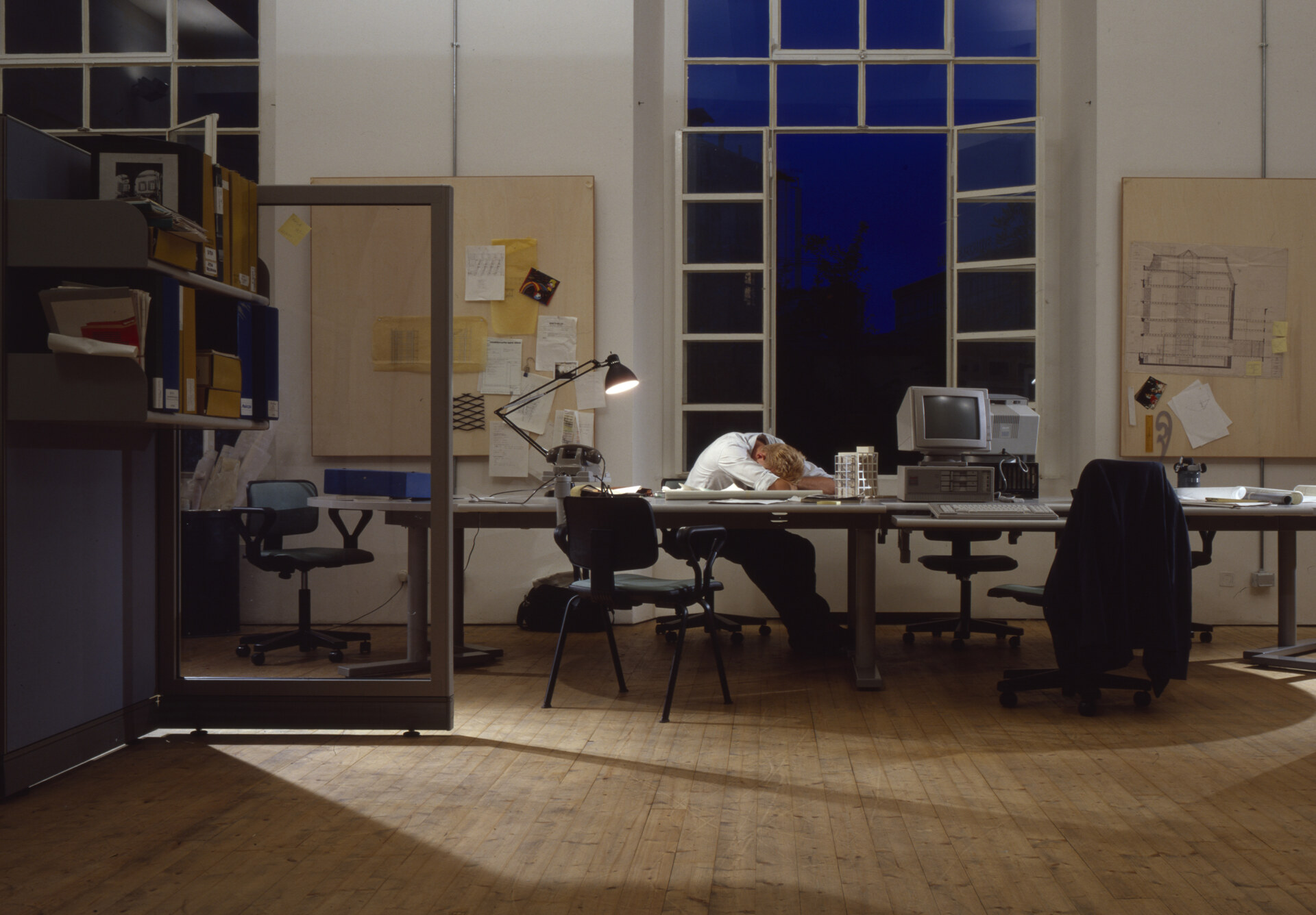 An Olivetti employee fast asleep by Santi Caleca - A person sleeps at a cluttered desk with a computer and lamp in a dimly lit office. The room has large windows showing a dark evening sky. Various papers and office furniture are scattered around.