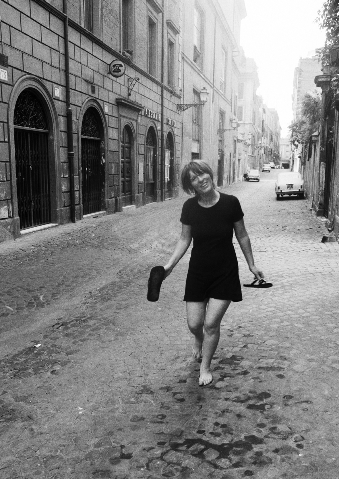 Photo by Santi Caleca, Letizia Battaglia in a black dress walks barefoot on a cobblestone street, holding shoes in one hand. Surrounding them are tall, old buildings with arched doorways and windows. A vintage car is parked further down the road, with trees and lamps lining the street.