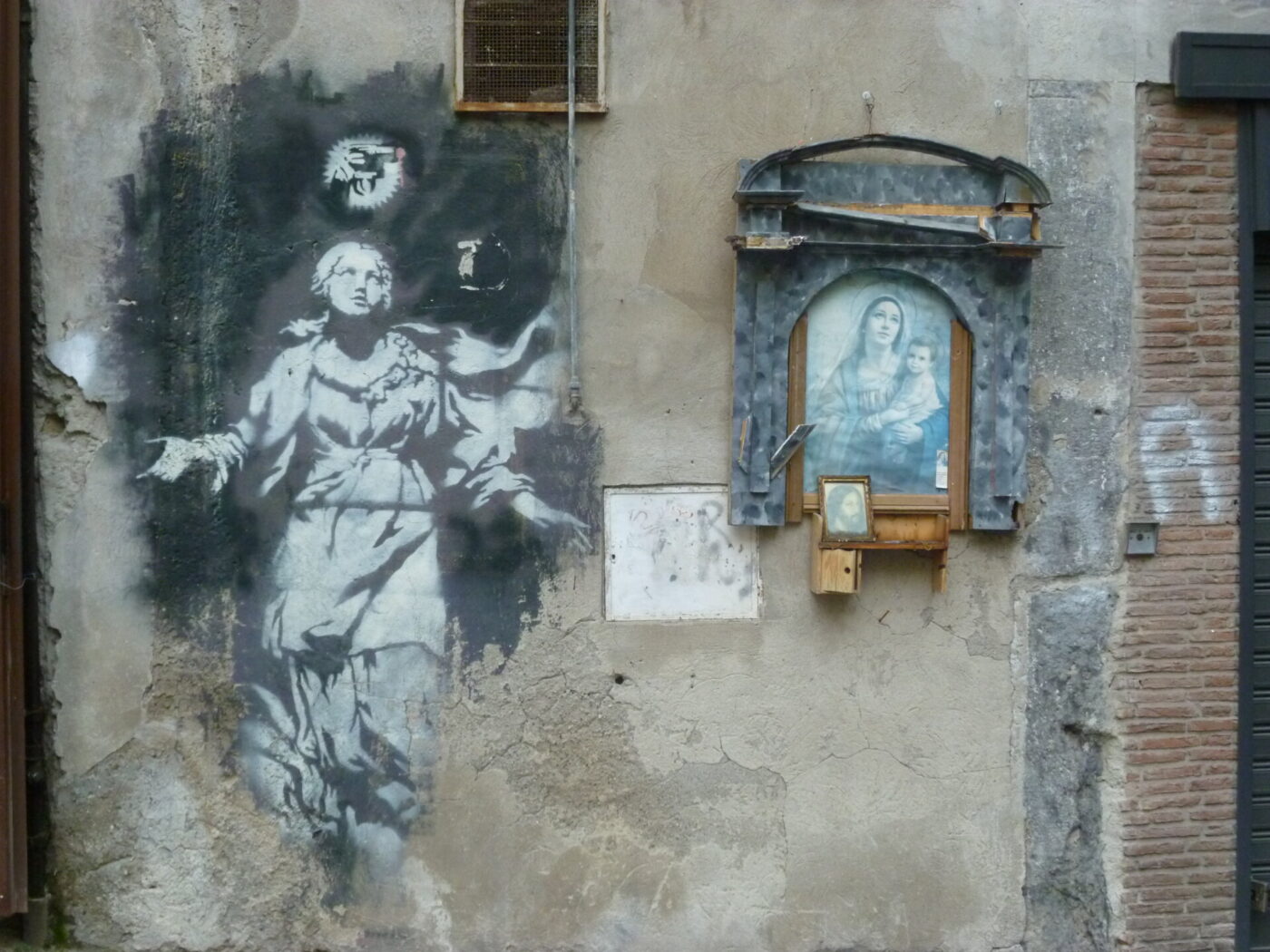Angel and Mary Shrine - Street art on a weathered wall depicts a monochrome angelic figure with hands outstretched. Beside it, a framed image of the Virgin Mary holding baby Jesus is displayed, housed in a small shrine with protective glass.