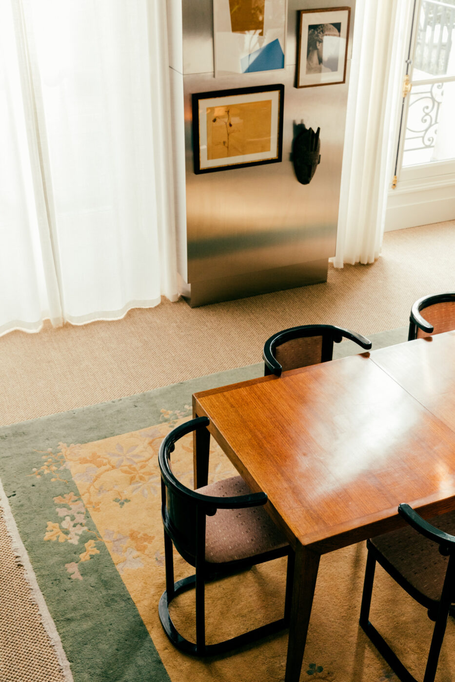 Un tavolo da pranzo in legno con quattro sedie con schienale curvo è sistemato in una stanza con grandi finestre coperte da tende bianche. Un'opera d'arte incorniciata e una maschera decorativa adornano un pannello metallico a parete. Il pavimento ha un tappeto con motivi floreali.
