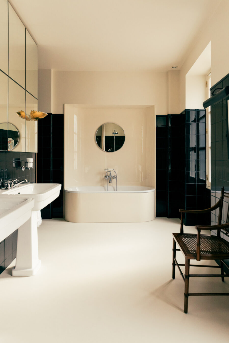 [Titolo dell'immagine] - Un bagno minimalista presenta una vasca da bagno bianca indipendente con uno specchio rotondo sopra. Le pareti sono piastrellate per metà con piastrelle scure. Un lavandino bianco è sulla sinistra e una sedia di legno è sulla destra. Lo spazio è ben illuminato dalla luce naturale.