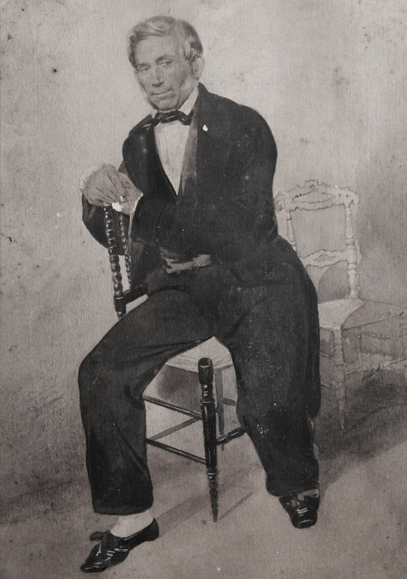 Early Chiavarina chair model - Black and white portrait of a man sitting on a wooden chair. He is dressed in formal 19th-century attire, including a dark suit and bow tie, resting one arm on the chair back and the other on his lap. Two empty chairs are in the background. If you have an image title to include, please replace [Image Title] with the appropriate text.