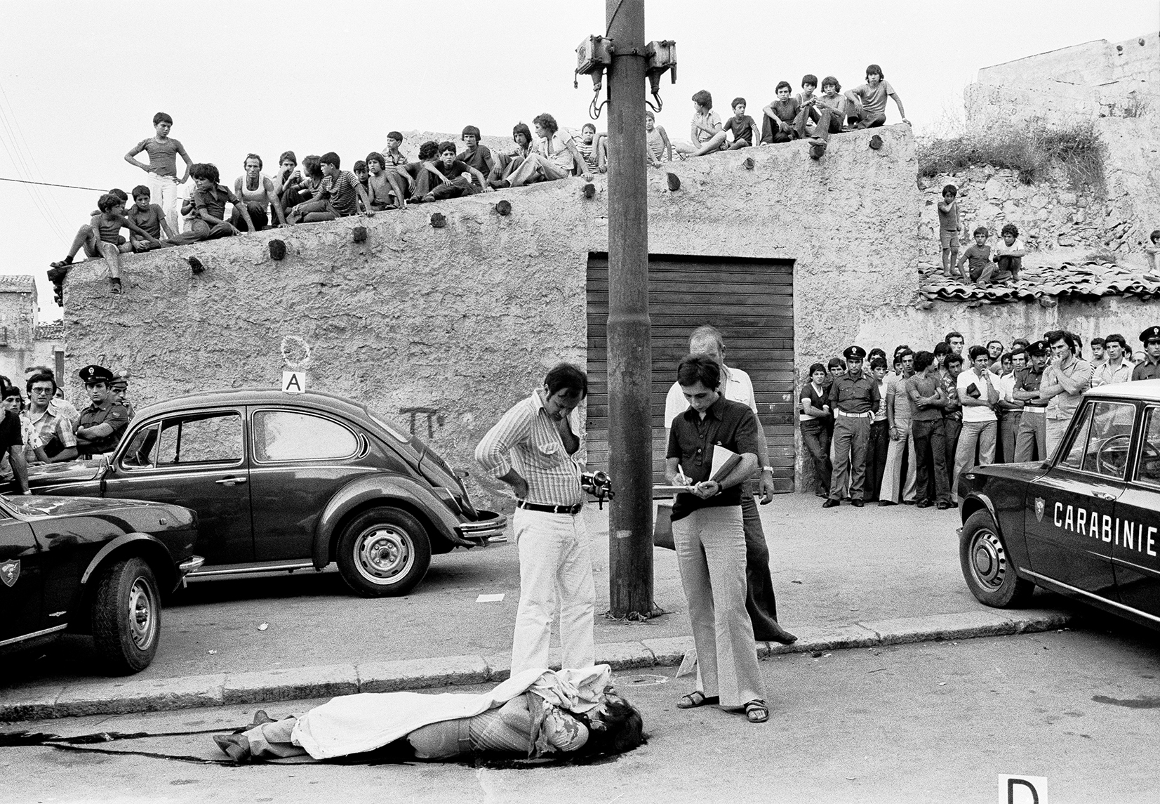 A murder in Palermo, 1976 (Santi used to cover mafia activity for the Palermitan left-wing daily L’Ora); - A group of people and police officers stand around a covered body on the street. Bystanders watch from a stone wall, and police cars are parked nearby. The scene appears tense and draws a crowds attention.