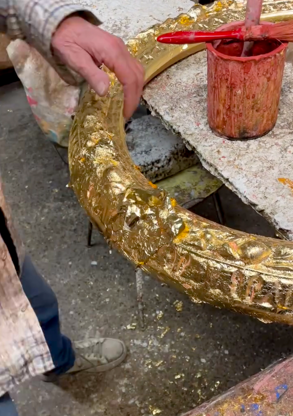 Giovanni Baccani : A person is holding and gilding an ornate, circular gold frame next to a table. The frame is partially covered in gold leaf. A red container with a brush is on the table. The person is wearing a plaid shirt and sneakers. Gold leaf fragments are on the floor.
