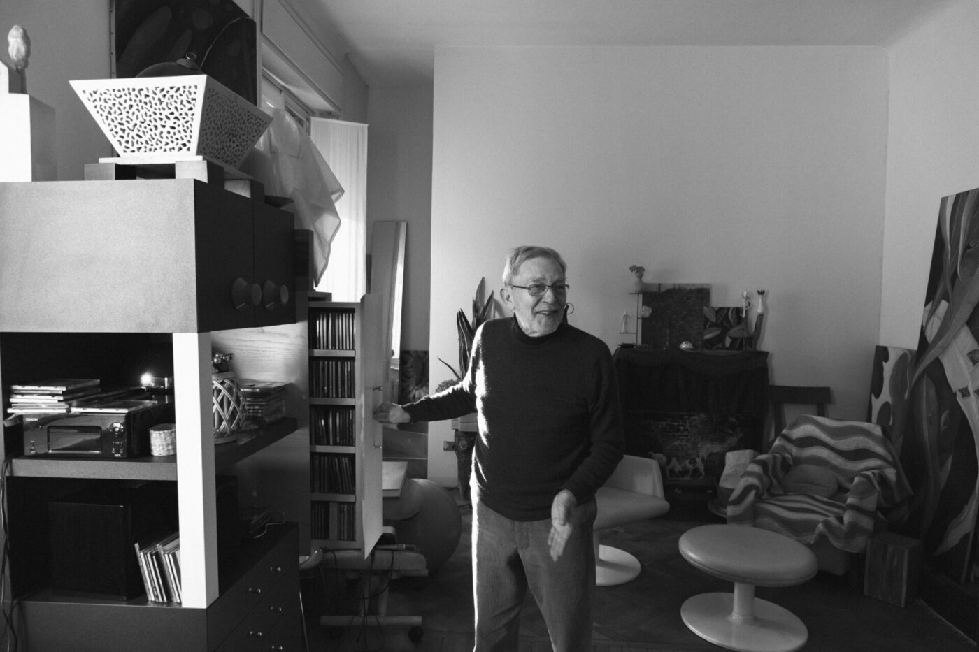 Santi Caleca stands in a well-decorated room, gesturing with his hands. The room contains modern furniture, a shelving unit with CDs, and abstract artwork. Sunlight softly illuminates the space. The mood is relaxed and inviting.