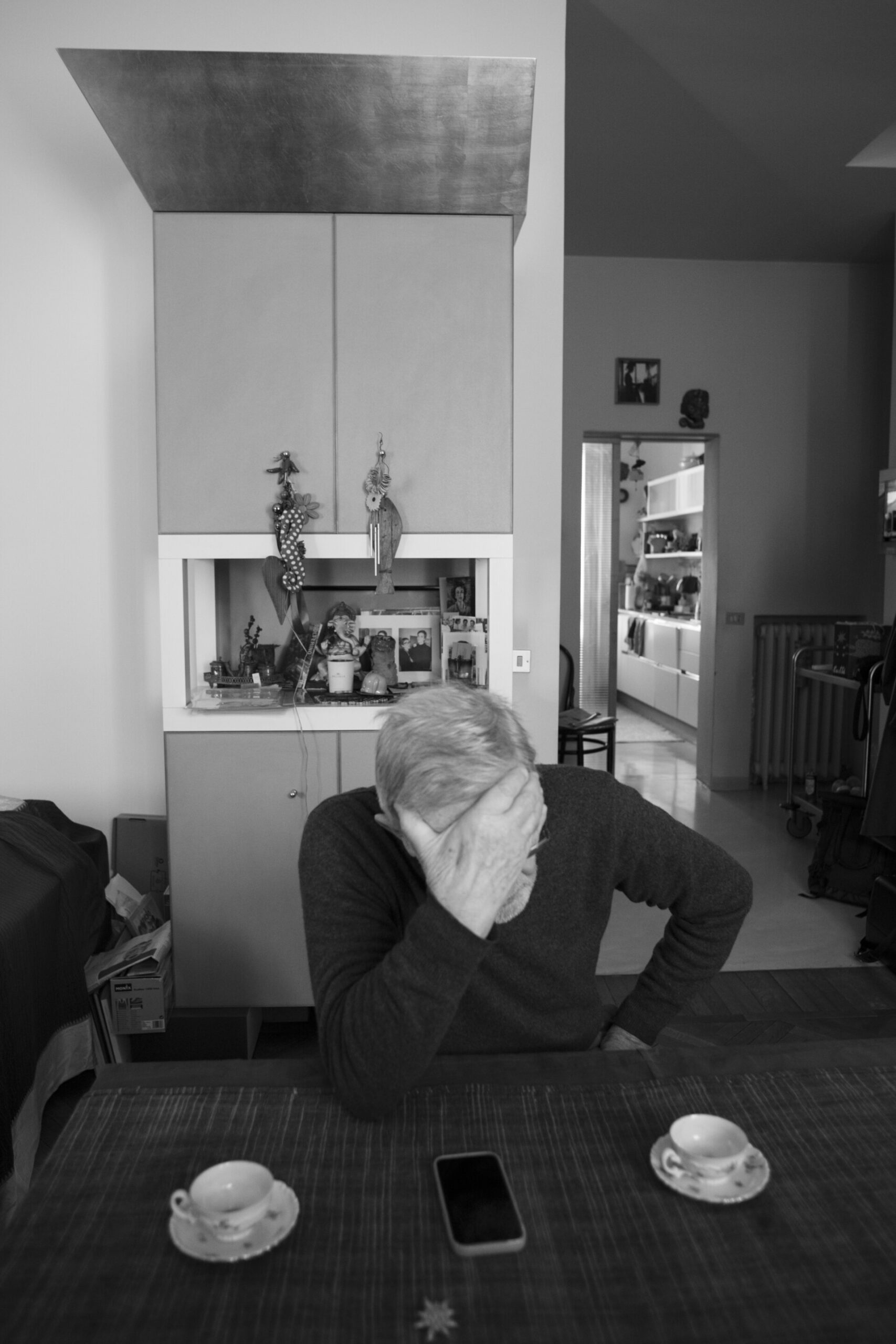 Santi Caleca - A person sits at a table with hands on their forehead, looking down. Two coffee cups and a smartphone are on the table. The background features a cabinet with decorations and a glimpse of a hallway. The image is in black and white.