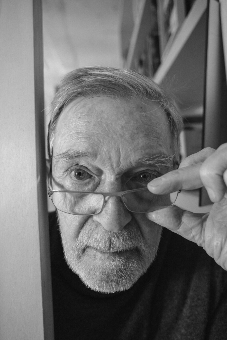 Focused Elder - An elderly man with a beard and glasses looks intently at the camera. He is adjusting his glasses with one hand, standing beside a bookshelf. The image is in black and white.