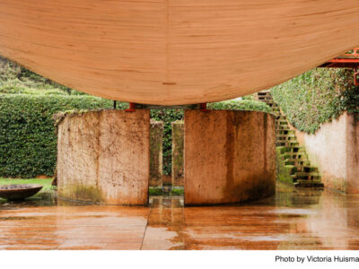 A curved wooden canopy with concrete pillars beneath it is part of a modern architectural structure. Surrounding greenery includes a neatly trimmed hedge and moss-covered stairs. The ground is wet, suggesting recent rainfall.