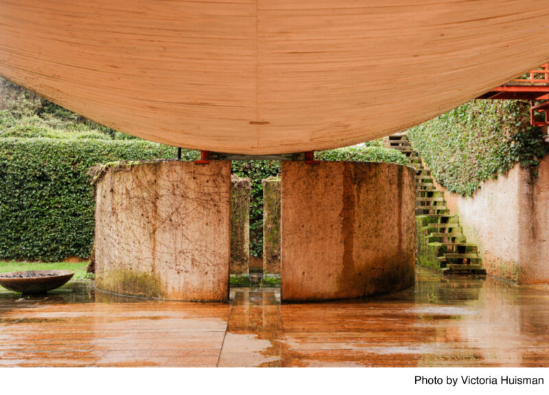 A curved wooden canopy with concrete pillars beneath it is part of a modern architectural structure. Surrounding greenery includes a neatly trimmed hedge and moss-covered stairs. The ground is wet, suggesting recent rainfall.