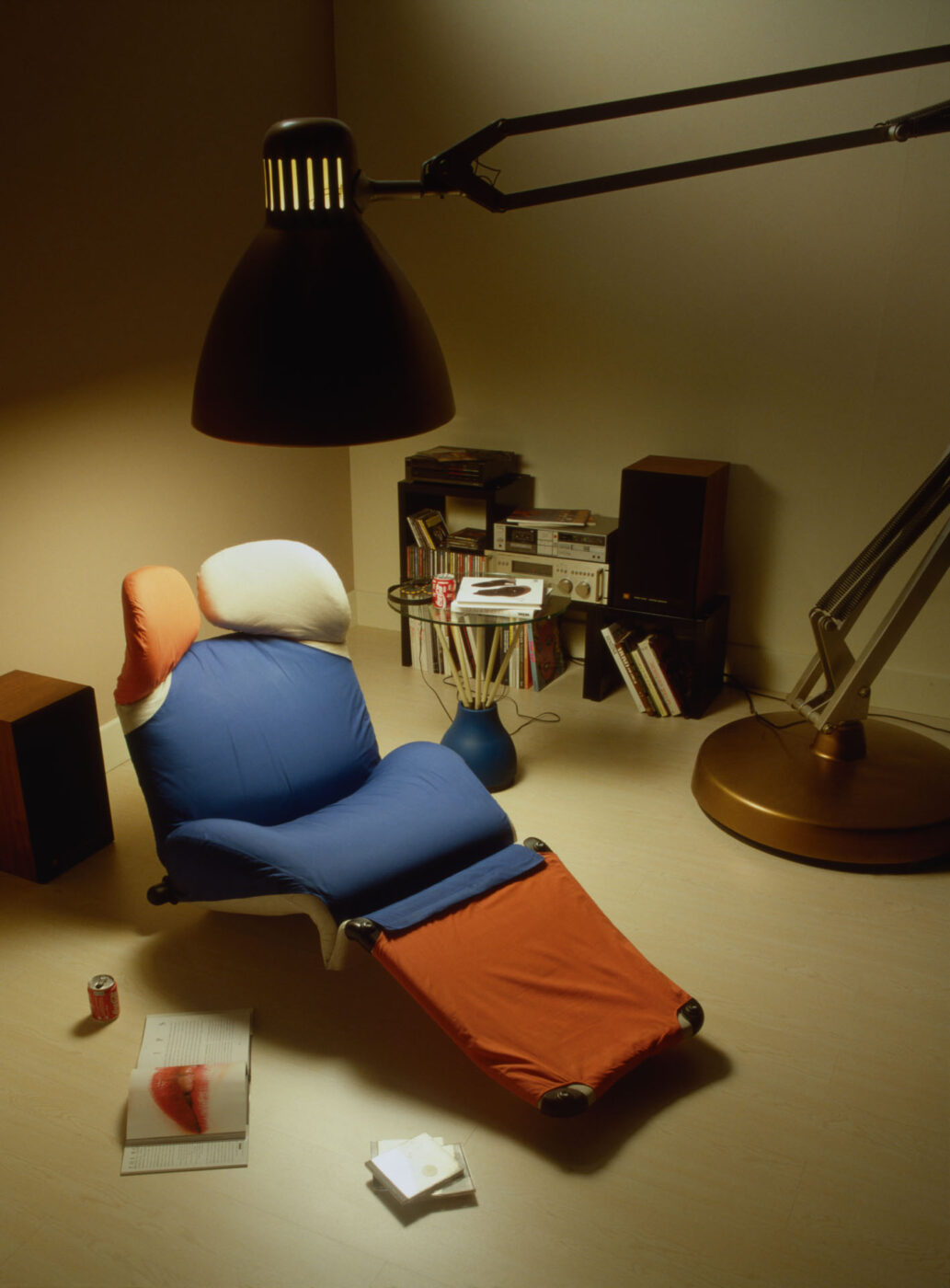 Cozy Reading Nook - A cozy room with a colorful recliner and a large overhead lamp. Books and decor fill a shelf in the corner, while magazines and a soft drink can are on the floor, creating a relaxed reading nook ambiance.
