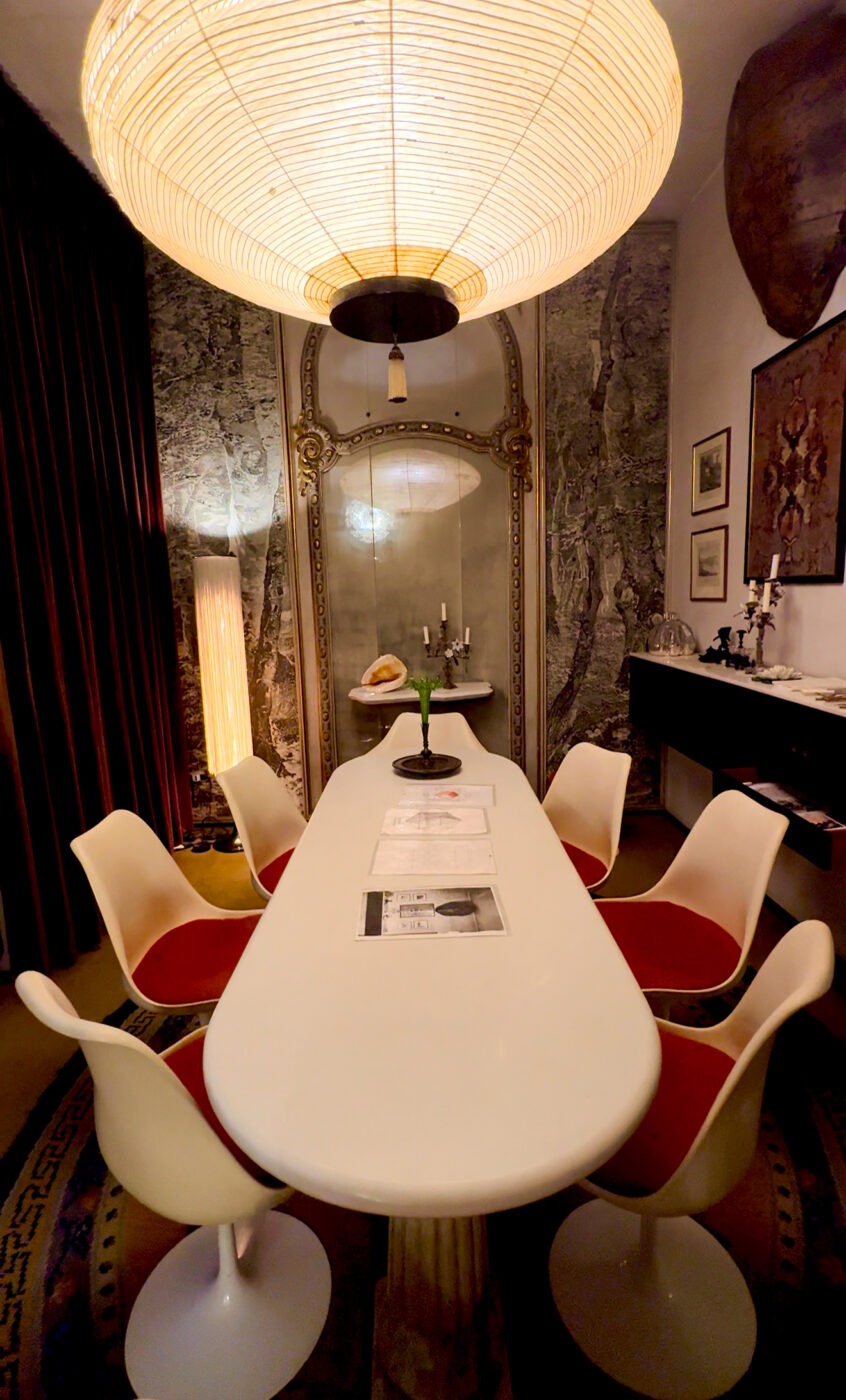 Modern Dining Room" - Una moderna sala da pranzo è caratterizzata da un lungo tavolo bianco circondato da sedie bianche con cuscini rossi. Una grande lampada rotonda sul soffitto fornisce un'illuminazione calda. Le pareti sono decorate con opere d'arte e specchi e una lampada alta e stretta è posta accanto a pesanti tende.